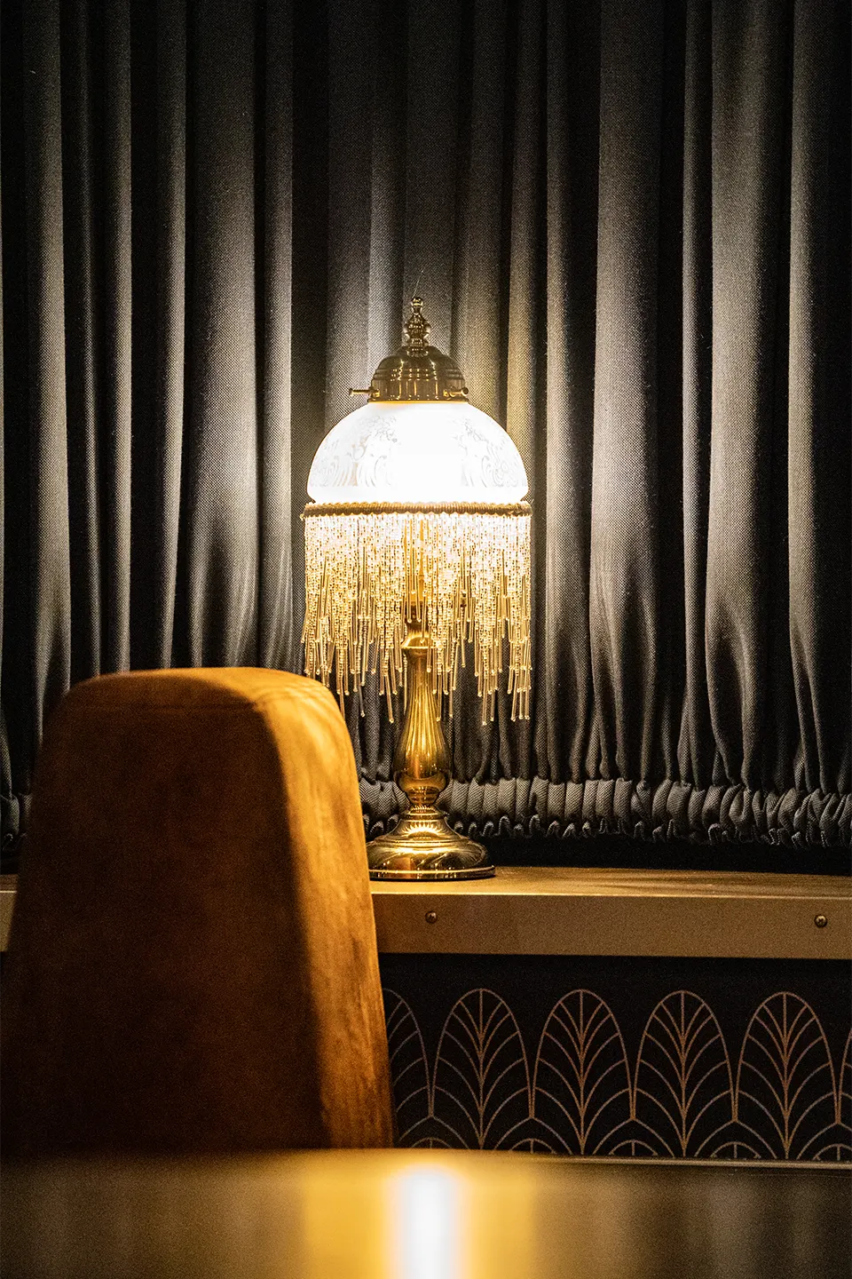 Goldene Tischlampe mit Fransen vor dunklem Vorhang neben einer samtbezogenen Sitzbank im stilvollen Art-déco-Ambiente.