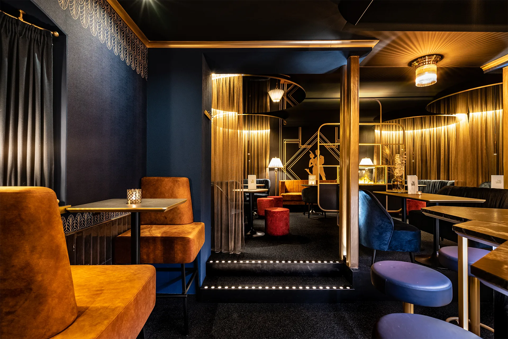 Ein eleganter Bar-Loungebereich im Art-Déco-Stil kombiniert dunkelblaue Wände mit goldenen Ornamenten und samtigen Sitzmöbeln in Orange, Blau und Rot.