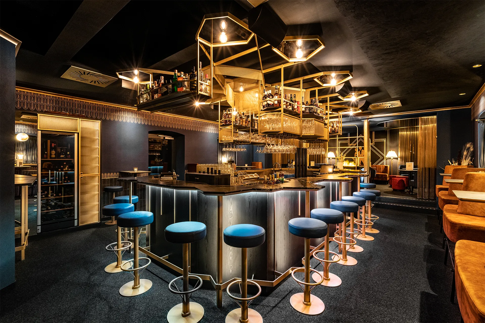 Eine polygonale, freistehende Bar mit dunkler Steinplatte und blauen Barhockern wird von einer auffälligen, geometrischen Regal-Konstruktion aus goldenem Metall gekrönt.