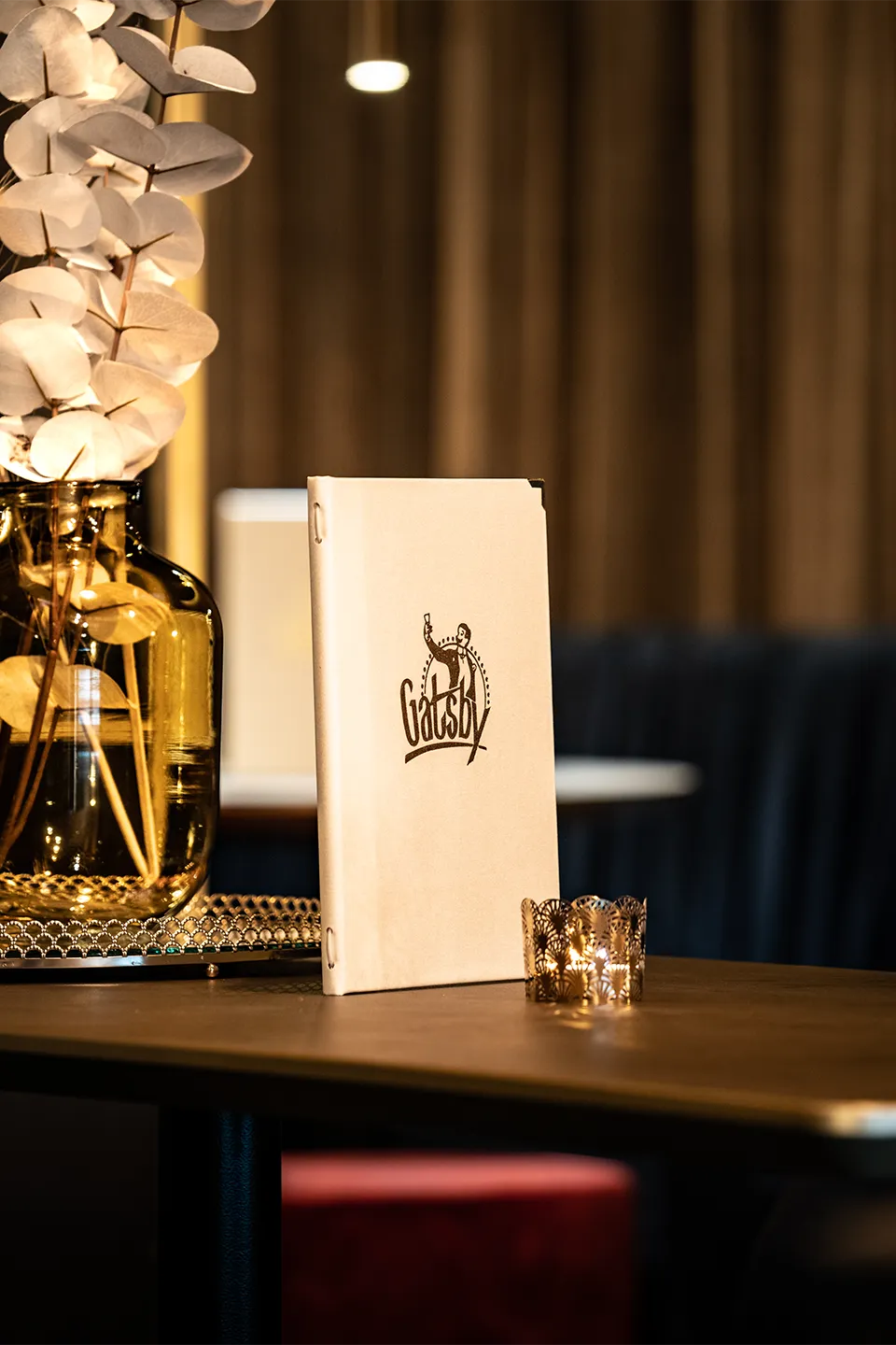 Auf einem dunklen Tisch steht eine weiße Menükarte mit dem schwarzen Logo „Gatsby“, dekoriert mit einem filigranen Teelichthalter und einer braunen Glasvase mit Eukalyptus.