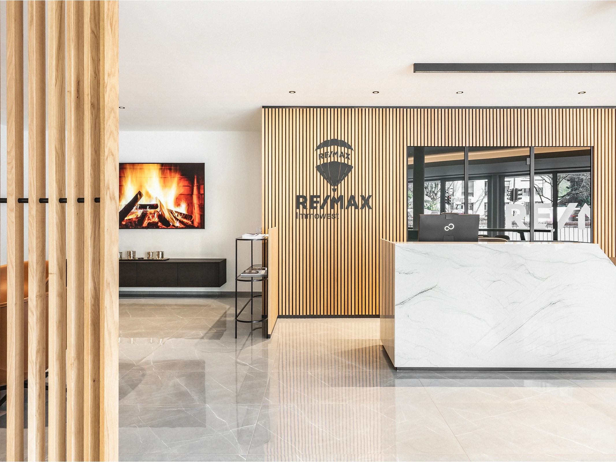 Empfangsbereich des RE/MAX Immowest Büros in Feldkirch mit weißem Marmortresen, hellem Holzlamellenregal, RE/MAX-Logo und Kaminbild im Hintergrund.