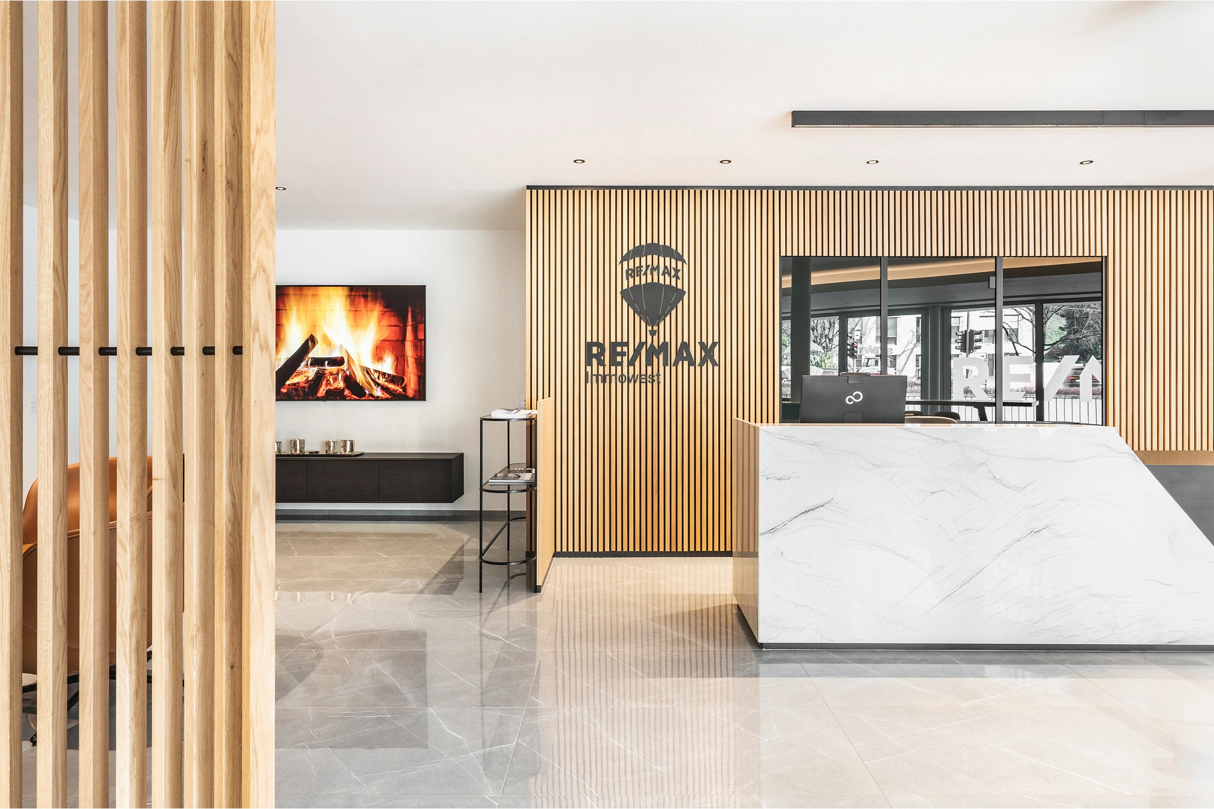 Empfangsbereich des RE/MAX Immowest Büros in Feldkirch mit weißem Marmortresen, hellem Holzlamellenregal, RE/MAX-Logo und Kaminbild im Hintergrund.
