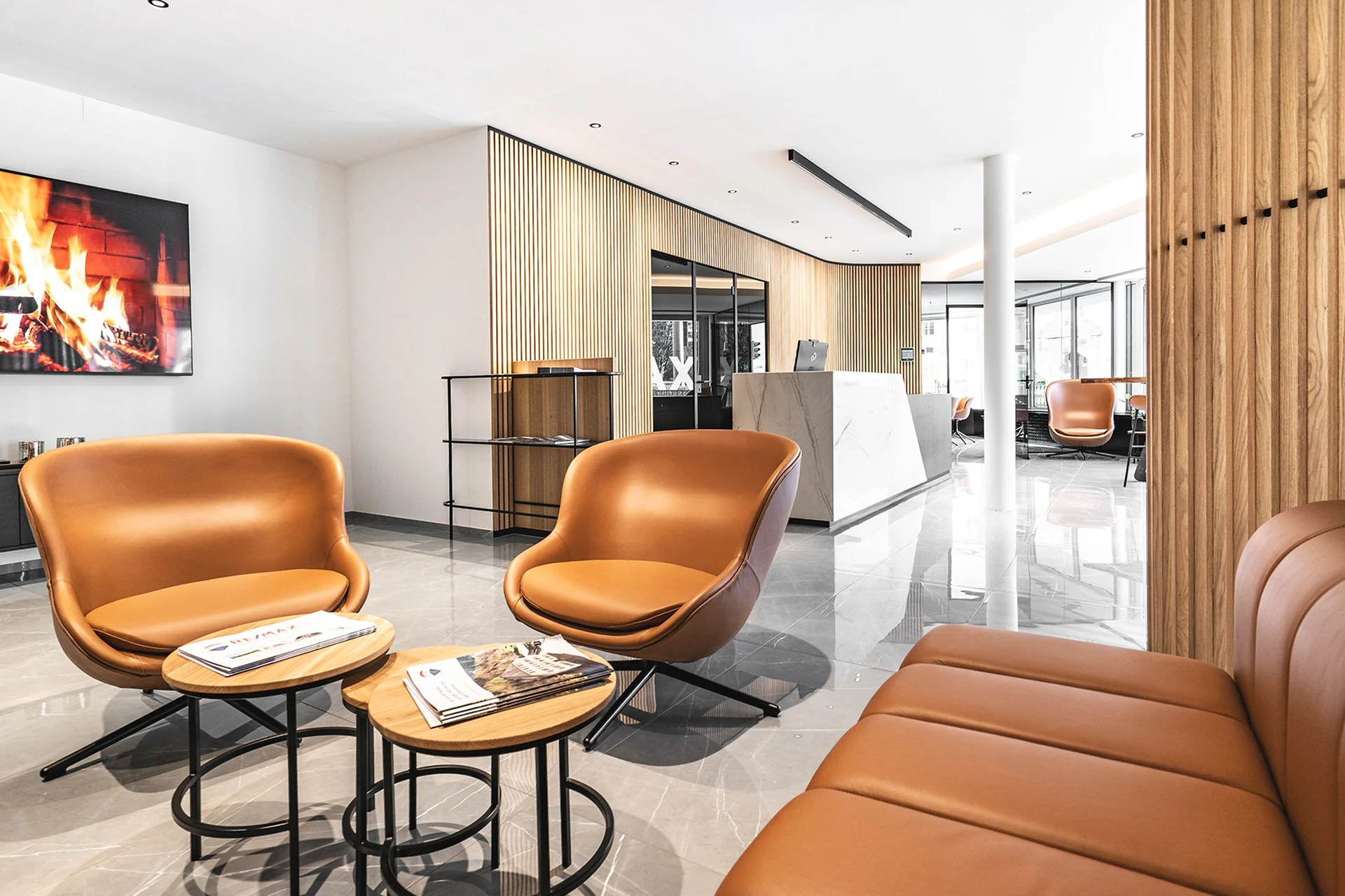 Wartebereich im RE/MAX Immowest Büro Feldkirch mit cognacfarbenen Ledersesseln, runden Holzcouchtischen, Kaminbild und Blick zur Marmorempfangstheke.