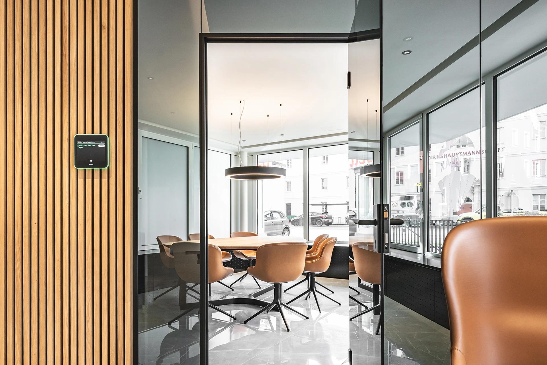 Blick durch die Glastür in den Besprechungsraum des RE/MAX Immowest Büros Feldkirch mit rundem Holztisch, cognacfarbenen Lederstühlen und ringförmiger Hängeleuchte.