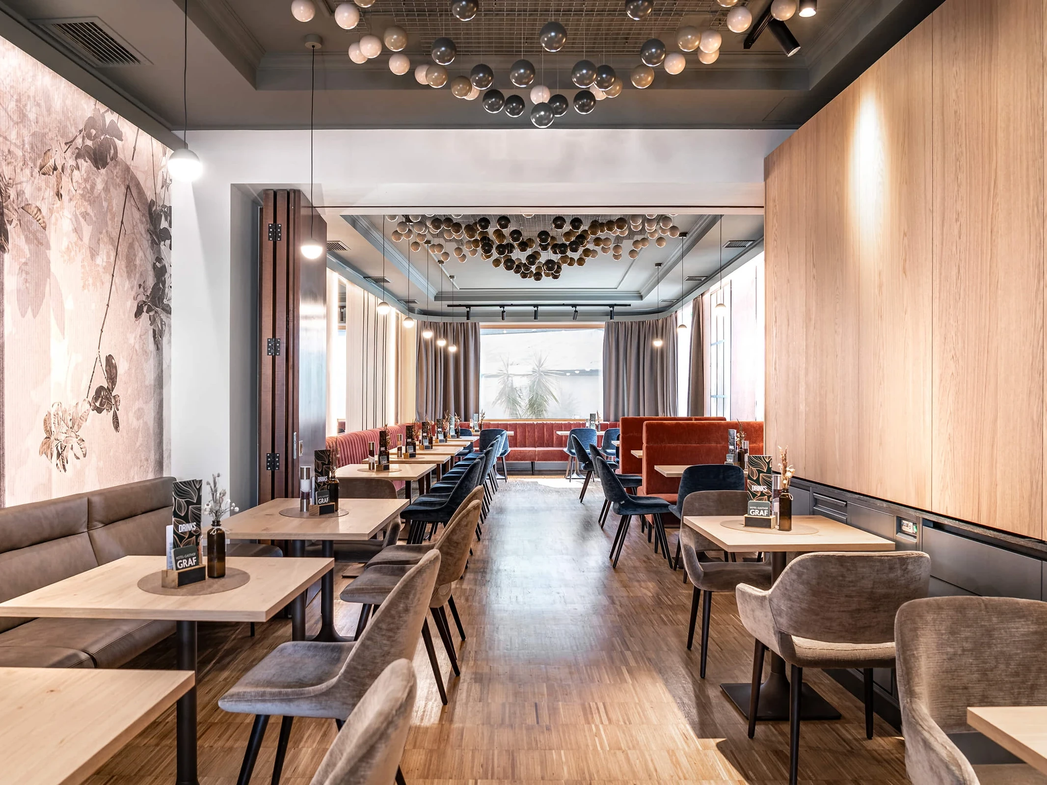 Weitläufiger Restaurantbereich im Hotel Graf St. Pölten mit hellen Holztischen, samtigen Stühlen in Grau und Blau, terrakottafarbenen Sitzbänken, floraler Wandtapete und einer markanten Deckeninstallation aus Glaskugeln.