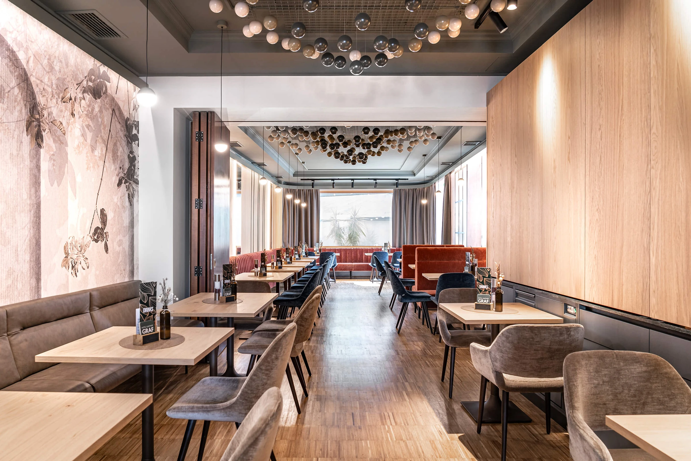 Weitläufiger Restaurantbereich im Hotel Graf St. Pölten mit hellen Holztischen, samtigen Stühlen in Grau und Blau, terrakottafarbenen Sitzbänken, floraler Wandtapete und einer markanten Deckeninstallation aus Glaskugeln.
