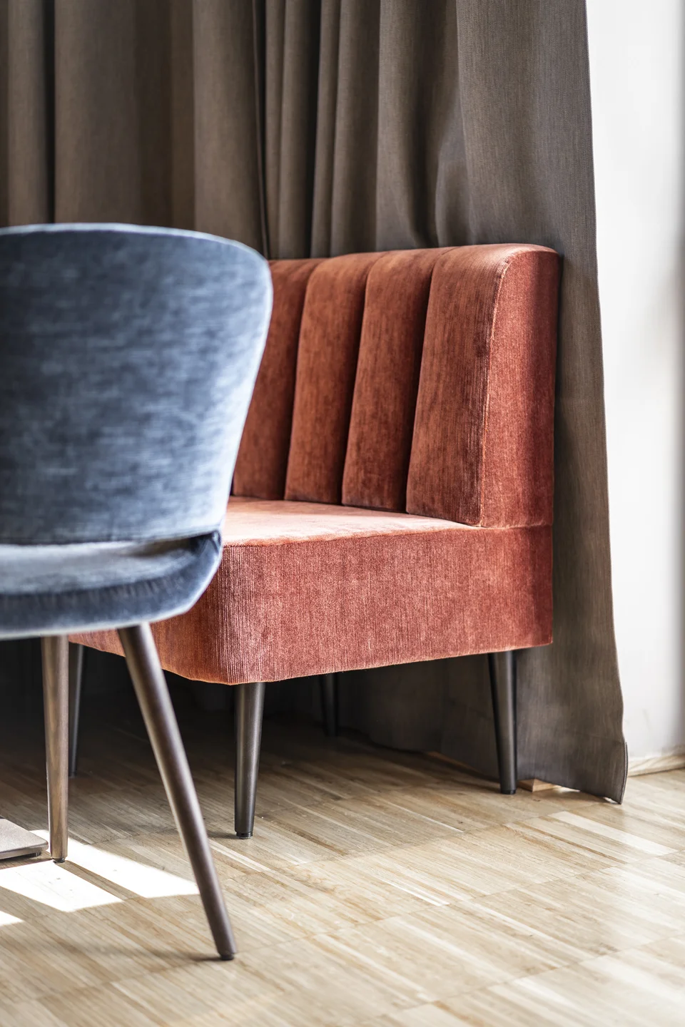 Detailaufnahme im Restaurant des Hotel Graf St. Pölten mit terrakottafarbenem Samtsessel mit Kanalsteppung, blauem Stuhl im Vordergrund und hellem Holzparkett vor graubraunem Vorhang.