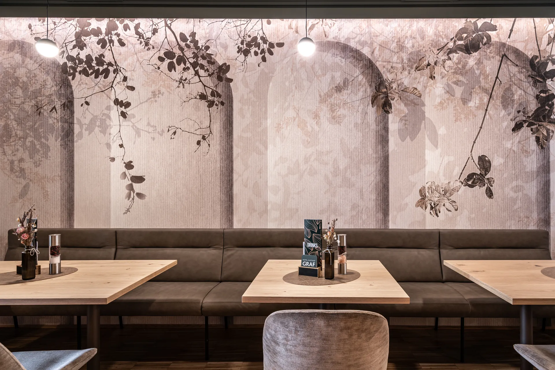 Sitzbereich im Restaurant des Hotel-Gasthof Graf St. Pölten mit langer Polsterbank in Graubraun, hellen Holztischen mit Trockenblumendekor und großformatiger floraler Tapete mit Blatt- und Bogenmotiv.