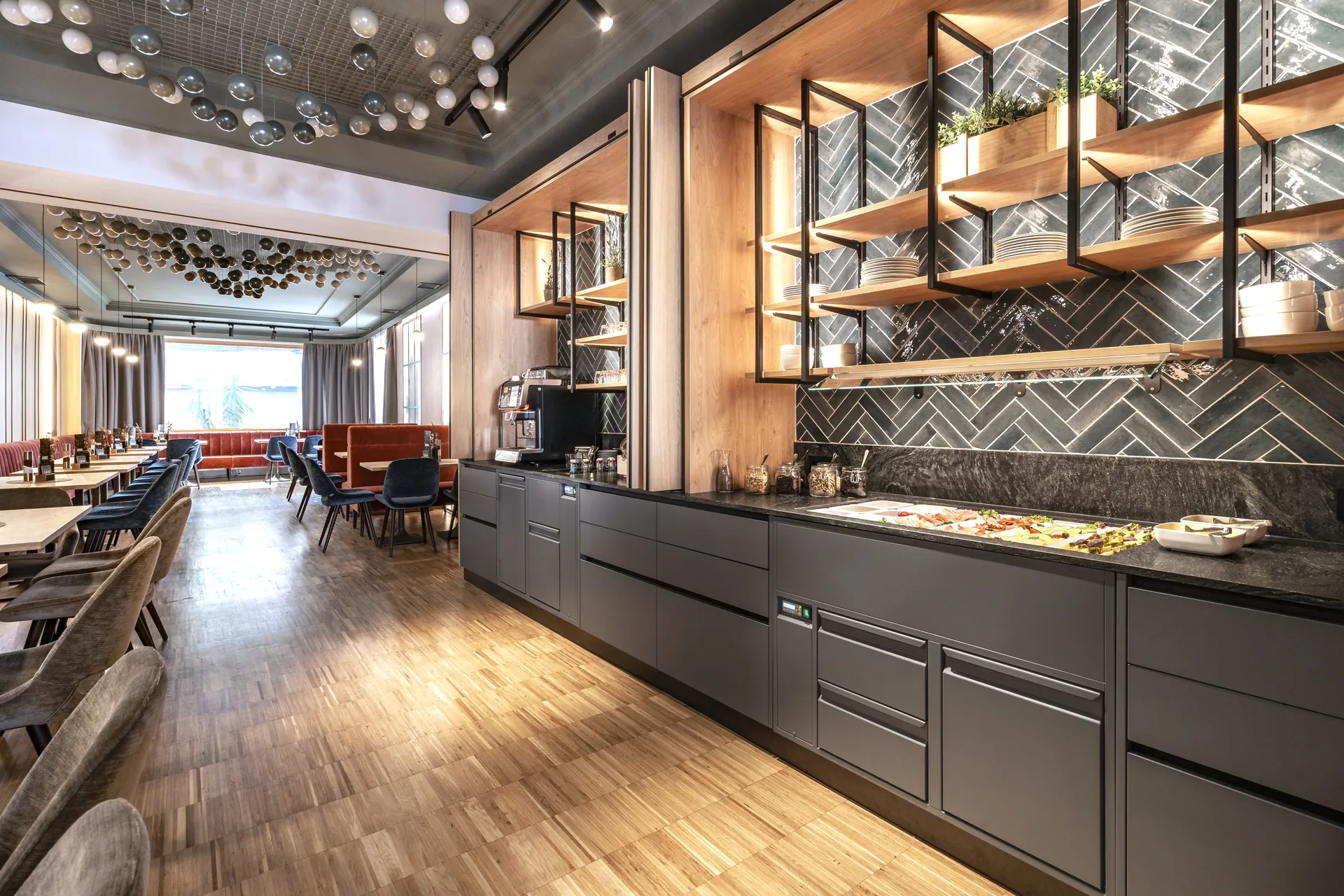 Blick in den Restaurantbereich des Hotel-Gasthof Graf St. Pölten mit Buffetzeile, offenen Holzregalen vor Fischgrät-Wandfliesen, Kugelleuchten-Deckeninstallation und dem langen Gastraum mit terrakottafarbenen Sitzbänken im Hintergrund.