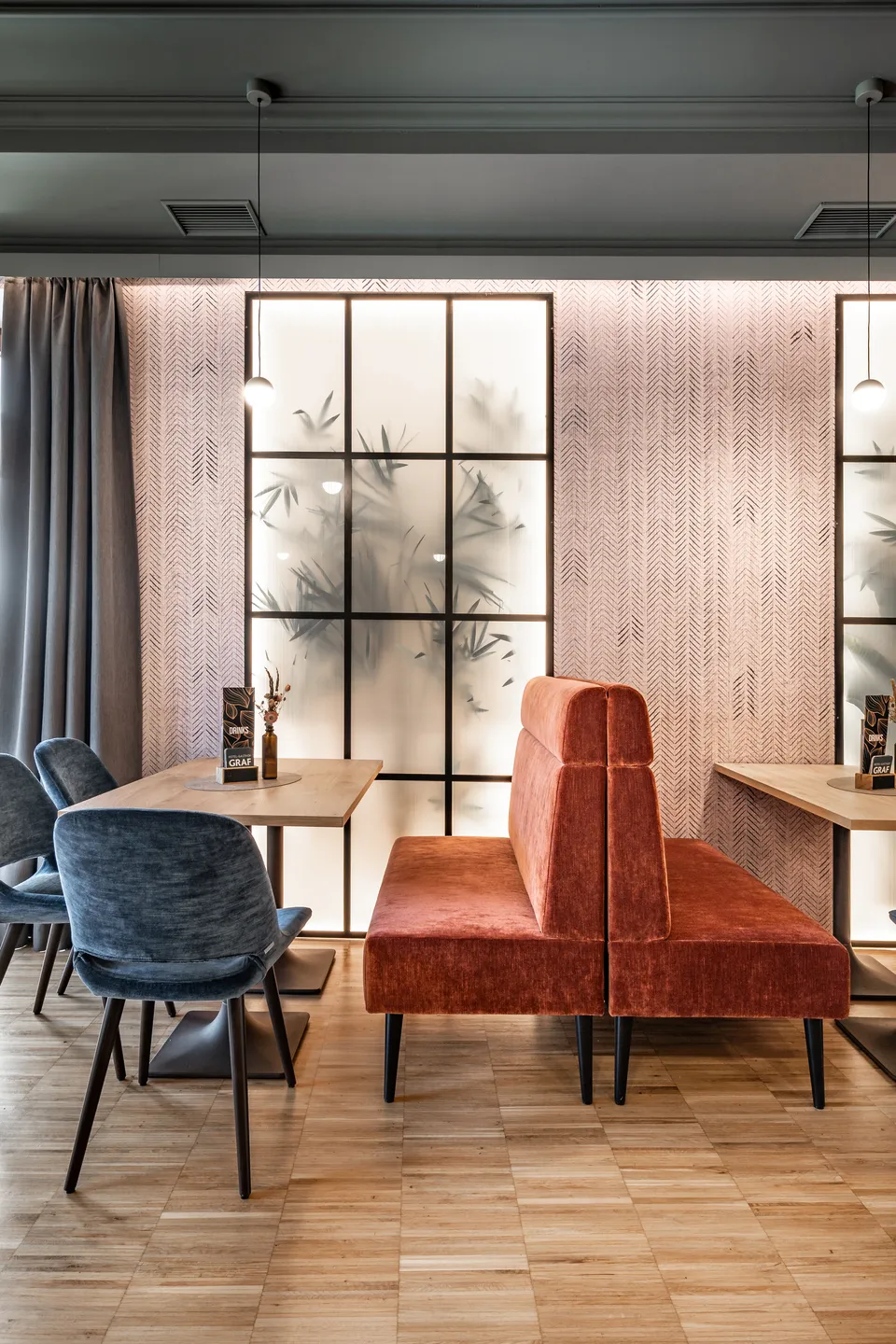 Restaurantbereich im Hotel-Gasthof Graf St. Pölten mit freistehendem terrakottafarbenem Samtsofa als Raumteiler, blauen Samtstühlen, Holztischen und hinterbeleuchtetem Fischgrät-Wandpaneel mit Bambus-Glaselement.