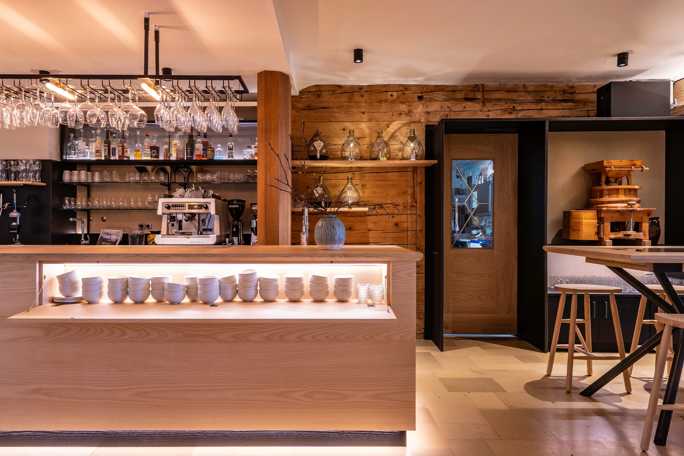 Warme Hotelbar mit hinterleuchteter Holztheke, aufgereihten Espressotassen, hängenden Weingläsern und einem Regal mit handgefüllten Schnapsballons vor rustikaler Altholzwand