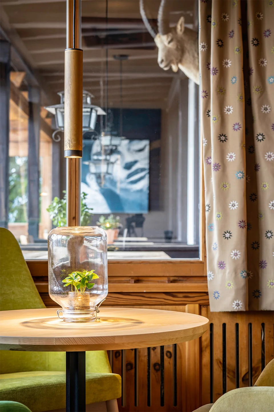 Gemütlicher Sitzplatz mit gelbgrünem Sessel, einer Pflanzenterrarium-Leuchte auf rundem Tisch und einem Vorhang mit Edelweißmuster vor einem Holzfenster