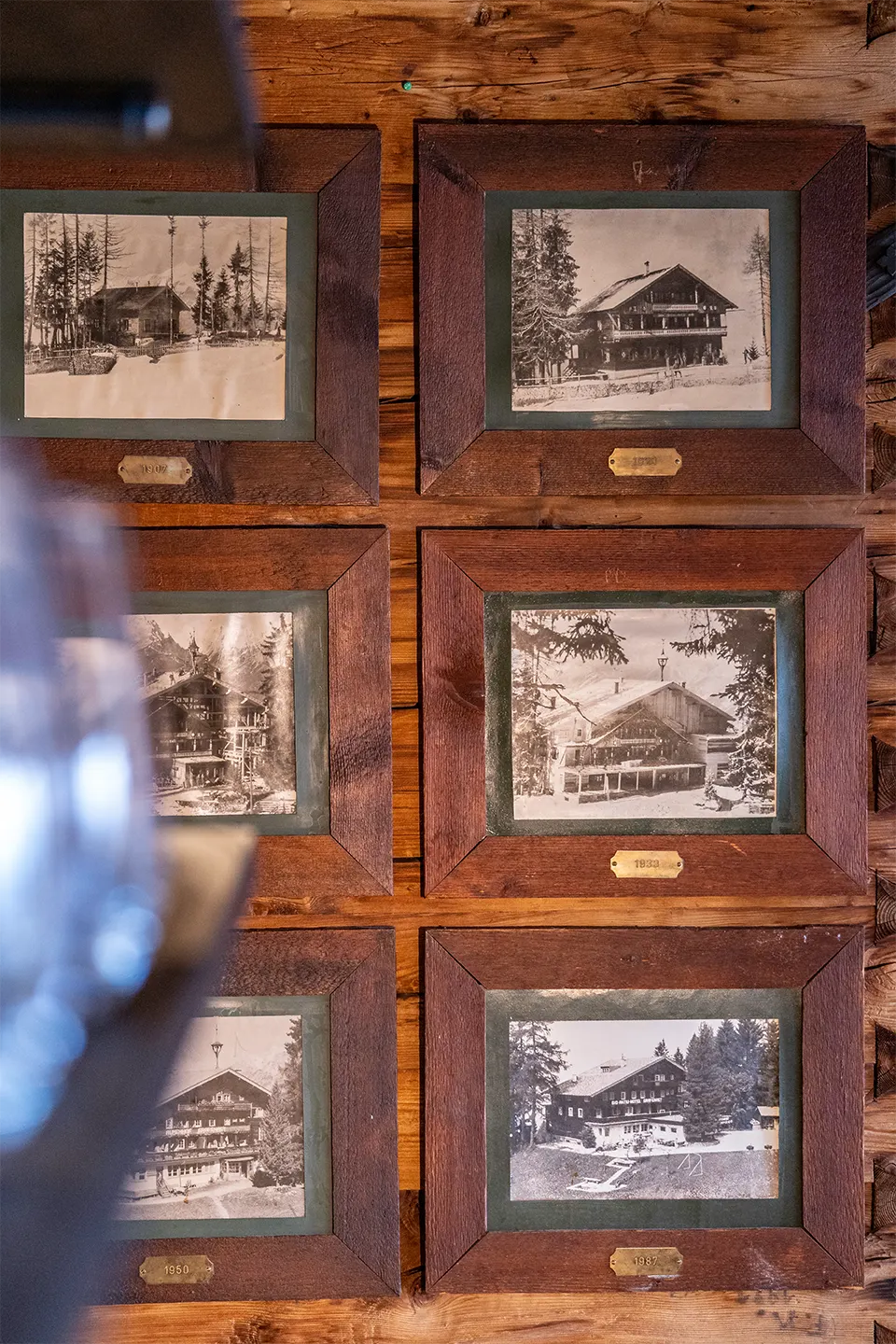 Historische Schwarz-Weiß-Fotografien des Alpenhofs aus verschiedenen Jahrzehnten, in dunkle Holzrahmen gefasst und auf einer Altholzwand arrangiert