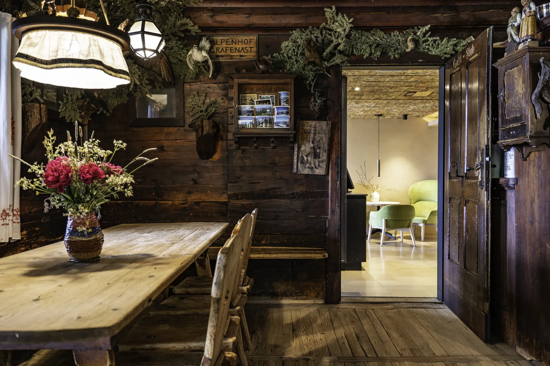 Rustikale Hotelstube mit dunkel gebeizter Holzvertäfelung, geschnitztem Türrahmen, frischem Blumenstrauß und Blick in den modern gestalteten Nachbarraum