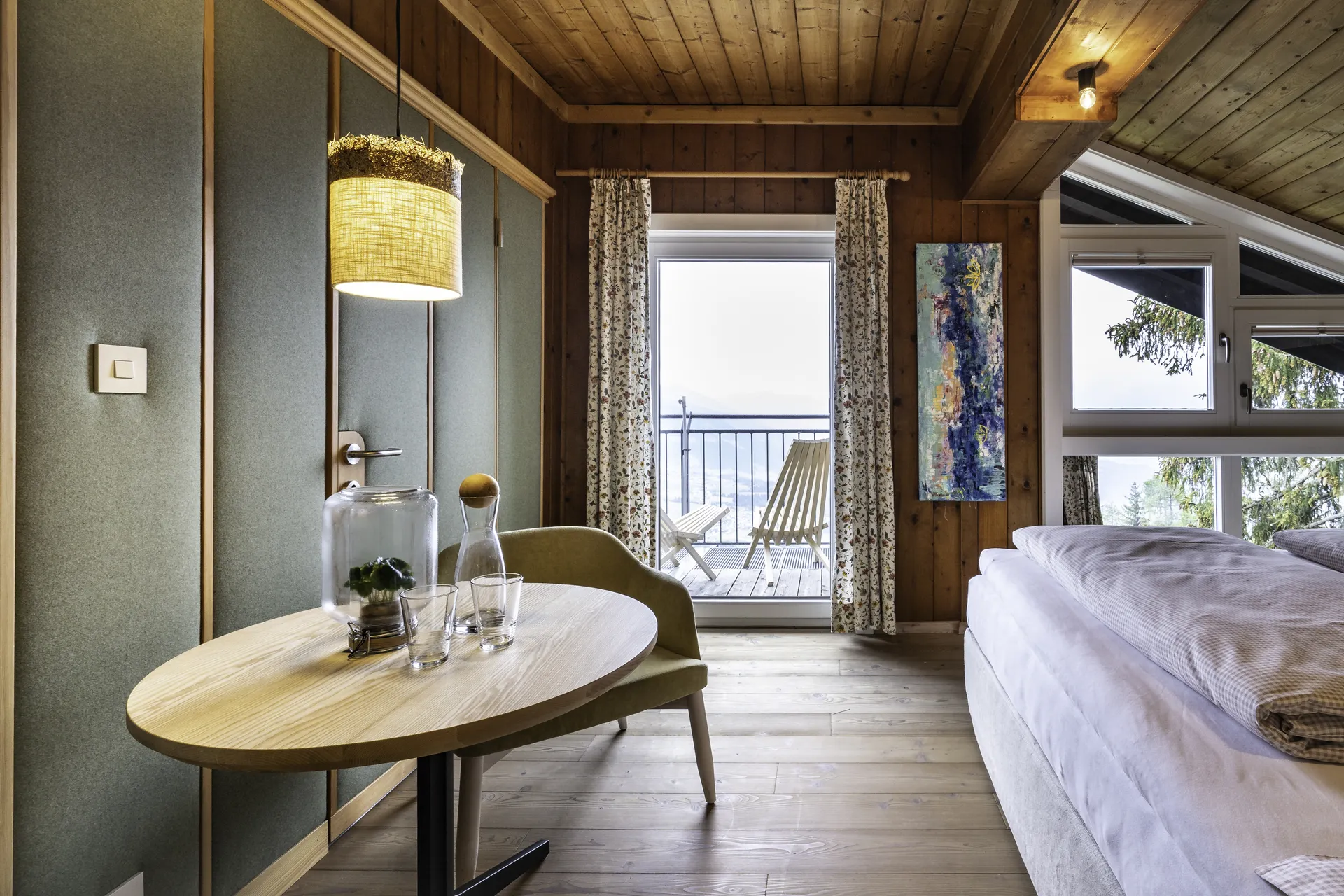 Alpines Hotelzimmer mit Holzdecke, grüner Stoffwand, geblümten Vorhängen und Balkonzugang mit Bergblick sowie einem modernen Sessel am Beistelltisch