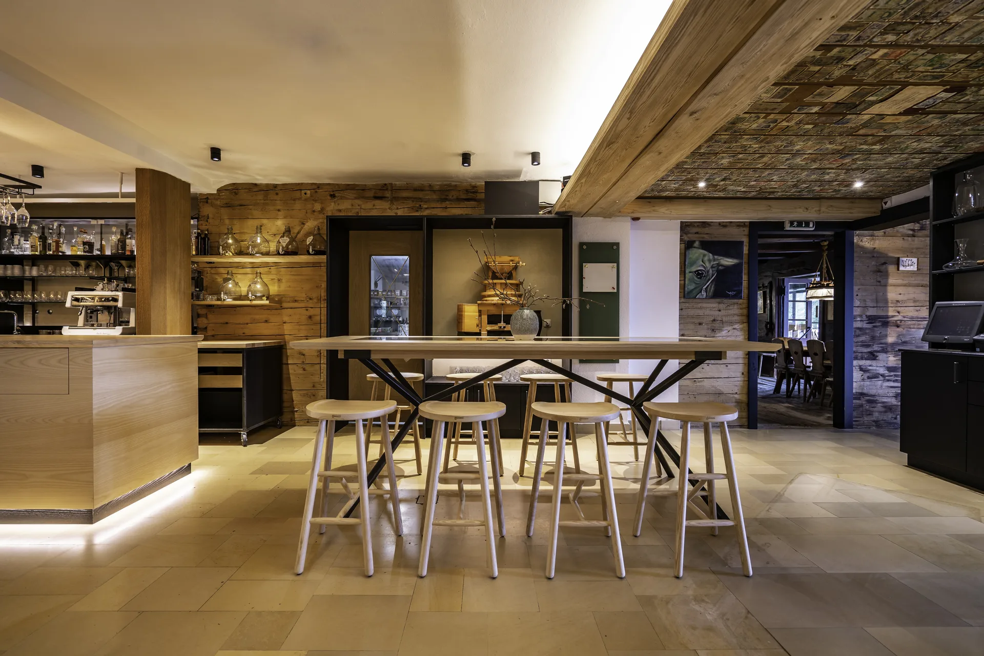 Zentrale Hotelbar mit langen Holztischen, hellen Barhockern, einem Altholz-Deckenbalken und Spirituosenregal vor einer warmen Altholzverkleidung