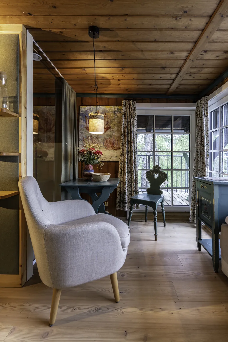 Gemütliches Hotelzimmer mit modernem Leinensessel, alpenländischem Herzstuhl, dunkelgrünem Schreibtisch, Ölgemälde und Balkonausblick in den Bergwald