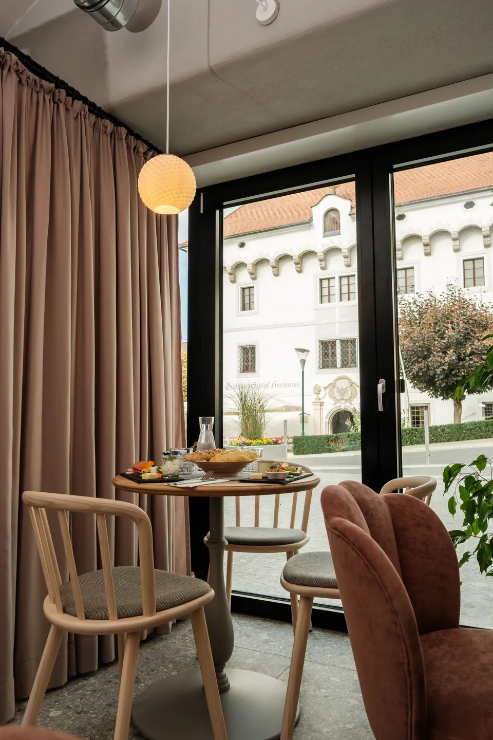 Cafébereich im Gwölb mit gedecktem Frühstückstisch, Holzstühlen, einer Kugelpendelleuchte und rosafarbenem Vorhang, durch das Panoramafenster Blick auf ein historisches Schlossgebäude.