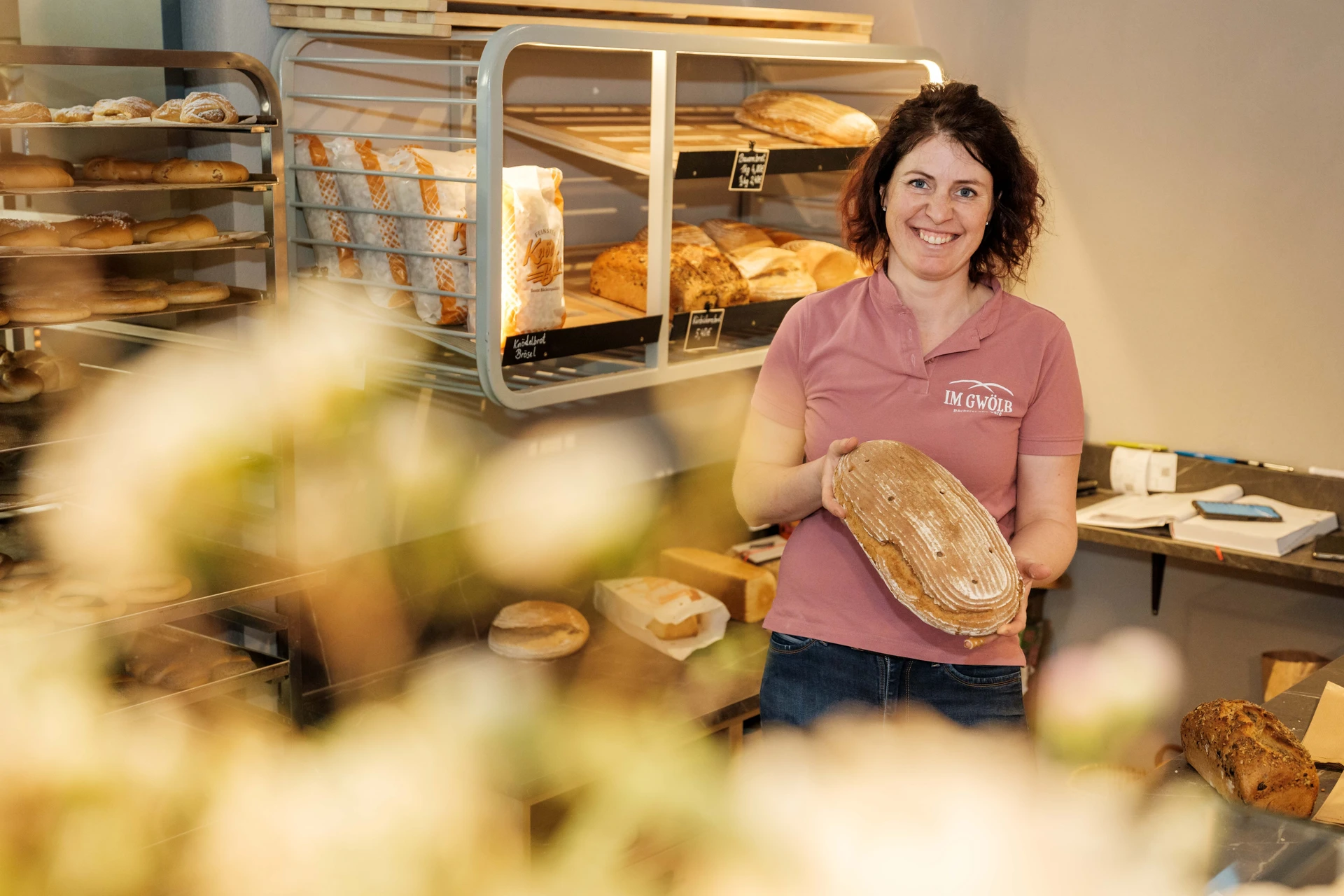 Inhaberin der Bäckerei im Gwölb mit einem frischen Sauerteigbrot in den Händen, lächelnd vor dem beleuchteten Brotregal mit verschiedenen Brotsorten.