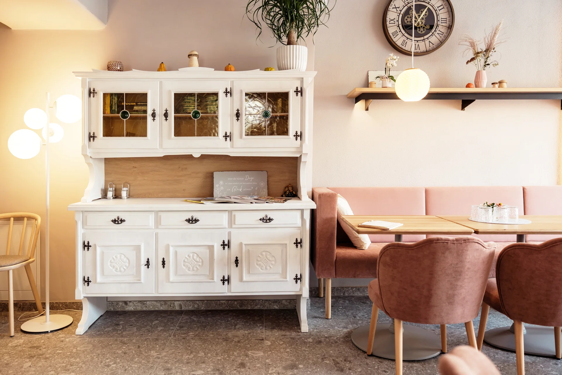 Cafébereich im Gwölb mit weißer antiker Kredenz mit Buntglasfenstern, rosafarbener Samtbank, herzförmigen Sesseln, hellen Holztischen, einer Kugelpendelleuchte und dekorativer Wanduhr.
