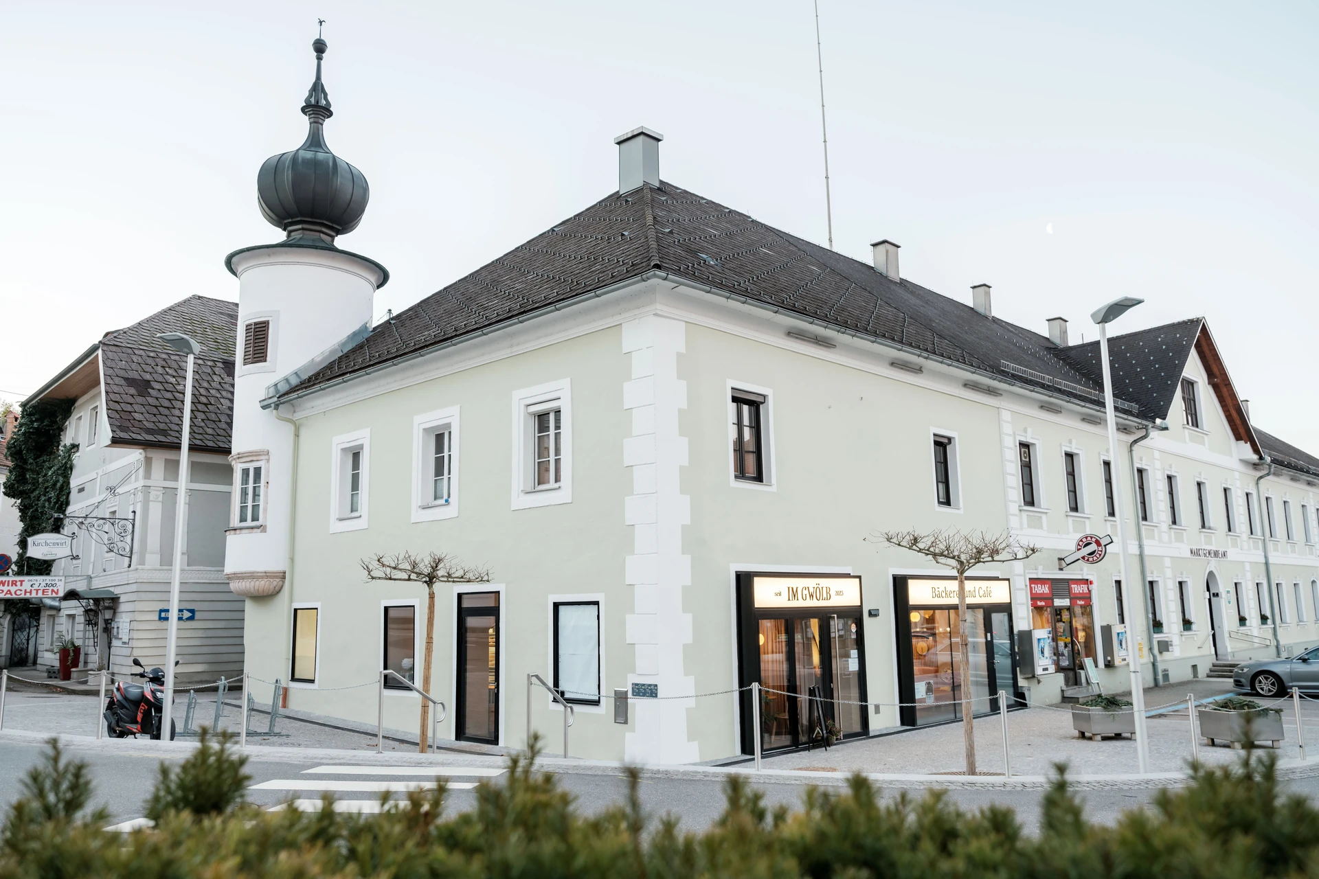 Außenansicht der Bäckerei und des Cafés Im Gwölb am Marktplatz mit historischem Gebäude, beleuchtetem Eingang und dem charakteristischen Zwiebelturm der Ortskapelle im Hintergrund.