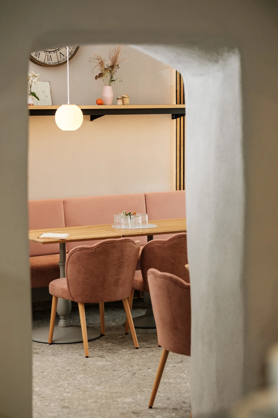 Blick durch einen historischen Gewölbebogen in den Cafébereich des Gwölb mit rosafarbener Samtbank, herzförmigen Sesseln, Holztisch, Kugelpendelleuchte und einem Wandregal mit Trockenblumen und Dekorationsobjekten.