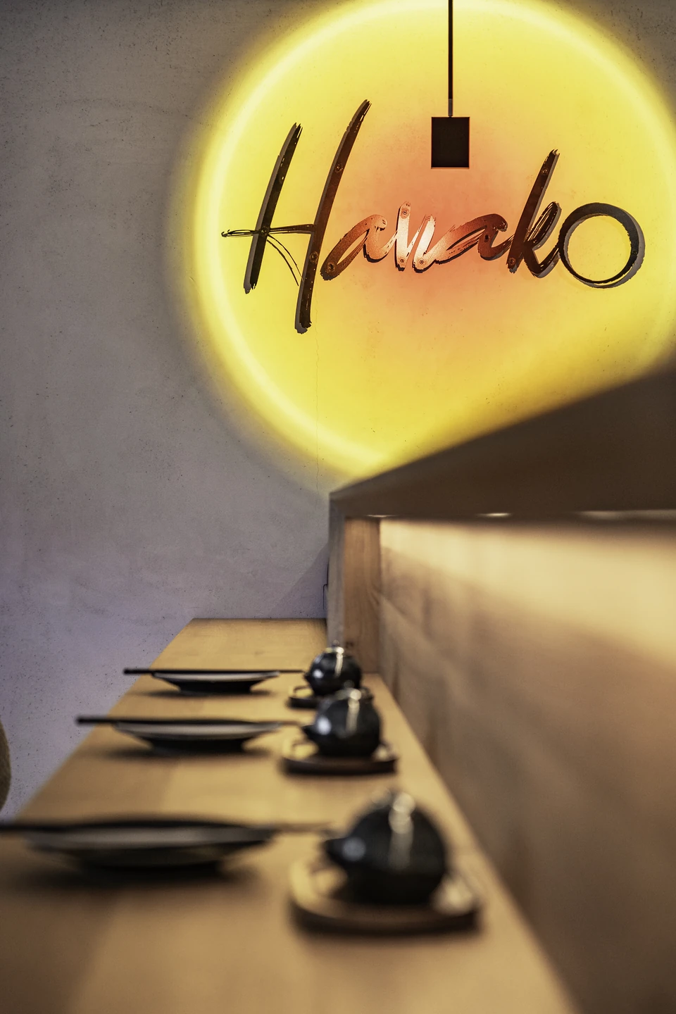Detail des beleuchteten Hanako-Schriftzugs an der Wand über einem gedeckten Tisch mit schwarzem Geschirr und Sitzbank in Dornbirn.