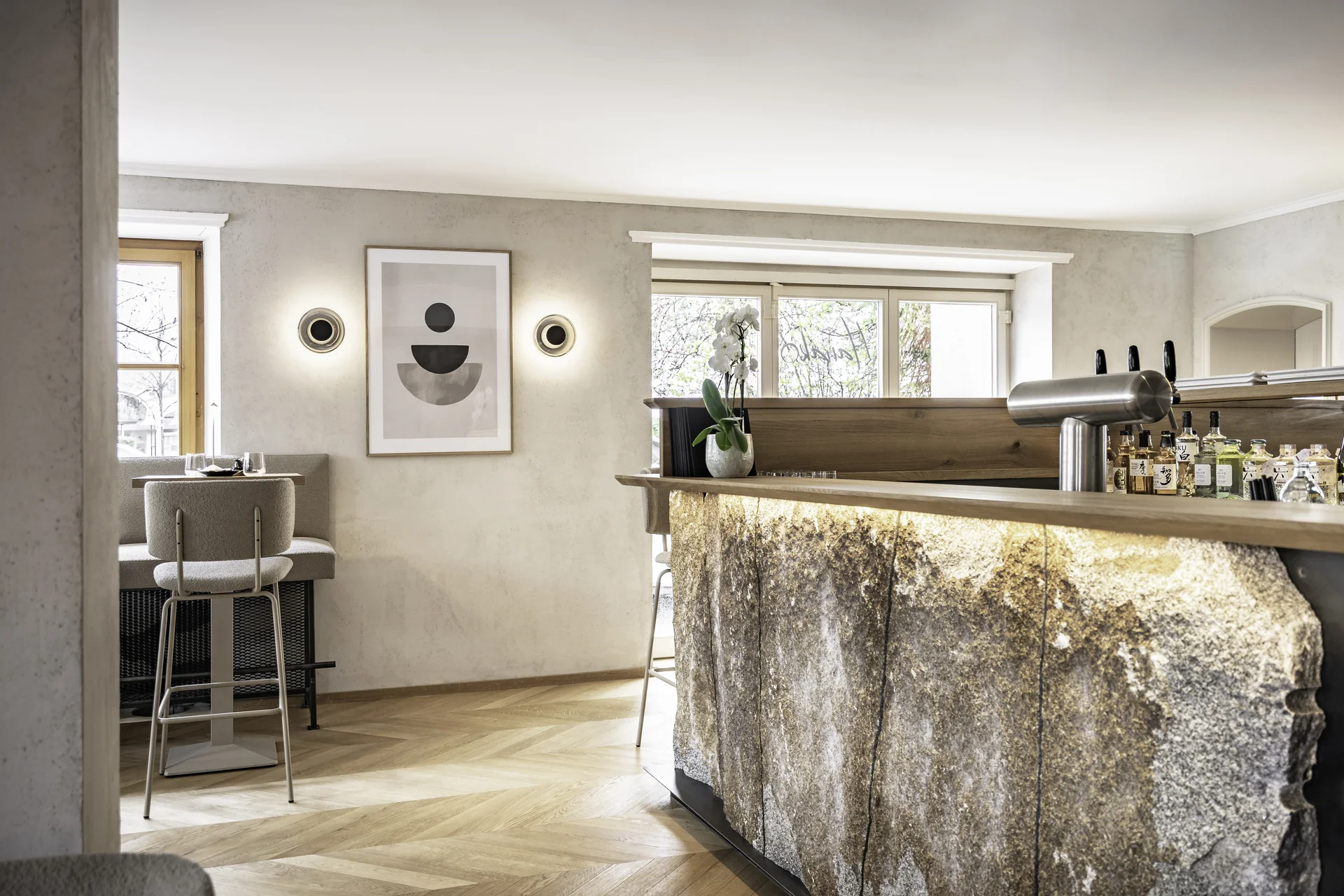 Bar im Hanako Dornbirn mit beleuchtetem Natursteintresen, Zapfanlage, Barhockern, abstraktem Wandbild, Wandleuchten und Fischgrätparkett.