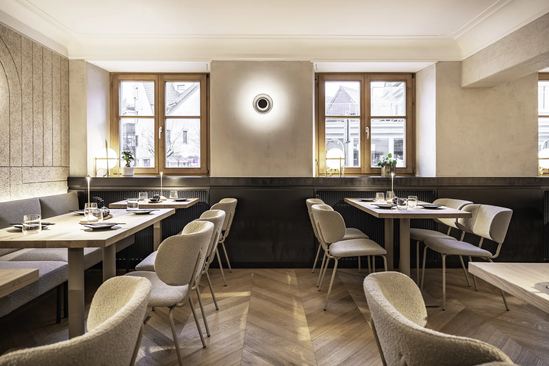 Gastraum im Hanako Dornbirn mit cremefarbenen Boucléstühlen, Sitzbank, gedeckten Holztischen, Fensterfront zur Straße, Wandleuchte und Fischgrätparkett.