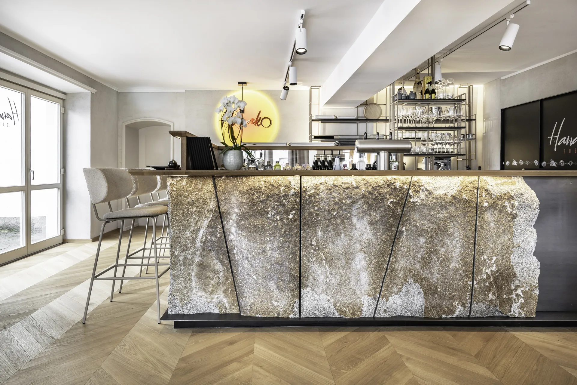 Bar im Hanako Dornbirn mit beleuchtetem Natursteintresen, Barhockern, Weinregal, Logo-Leuchtreklame und Fischgrätparkett.