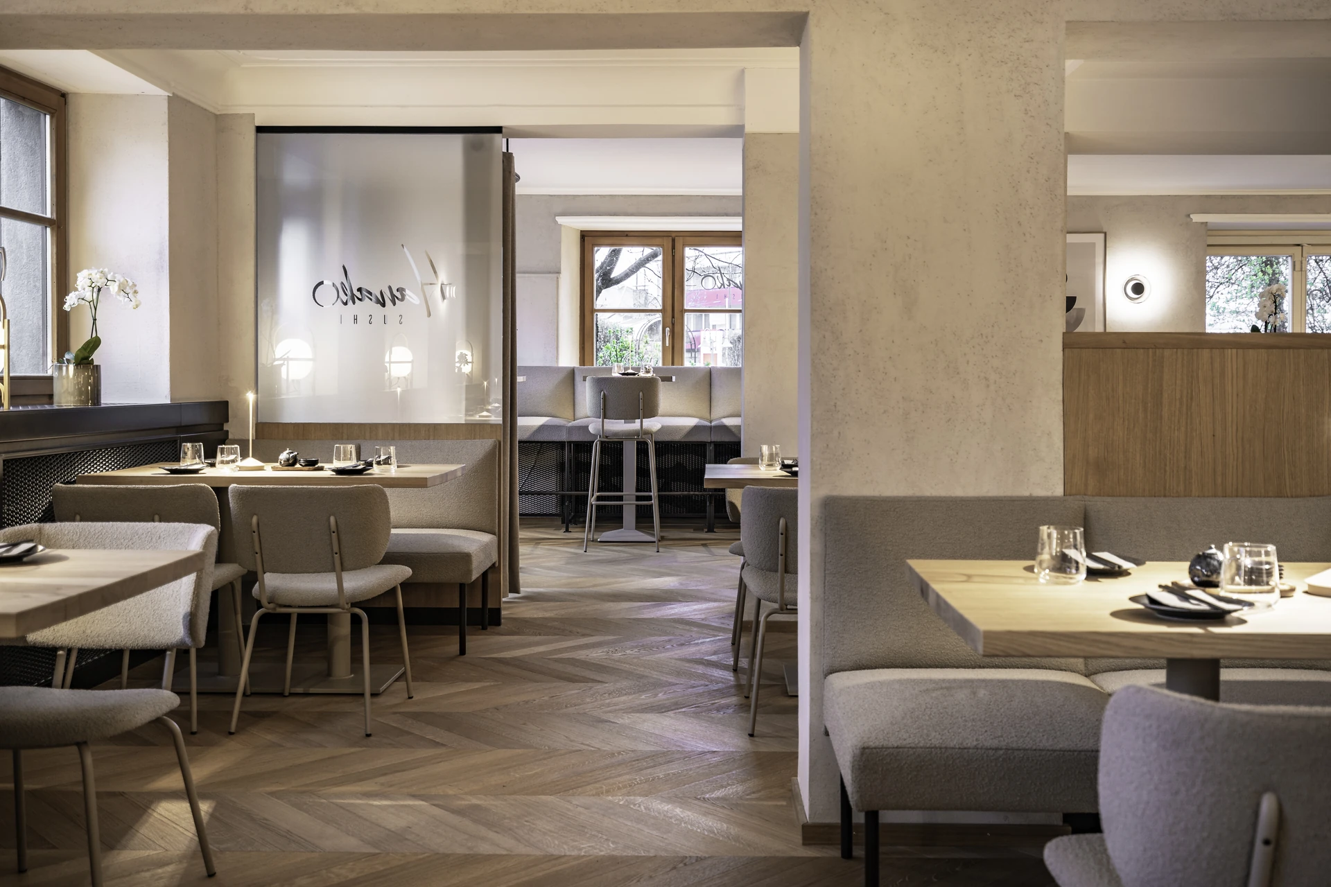 Gastraum im Hanako Dornbirn mit grauen Sesseln, Sitzbänken, gedeckten Holztischen, Glastrennwand mit Logo und Fischgrätparkett.