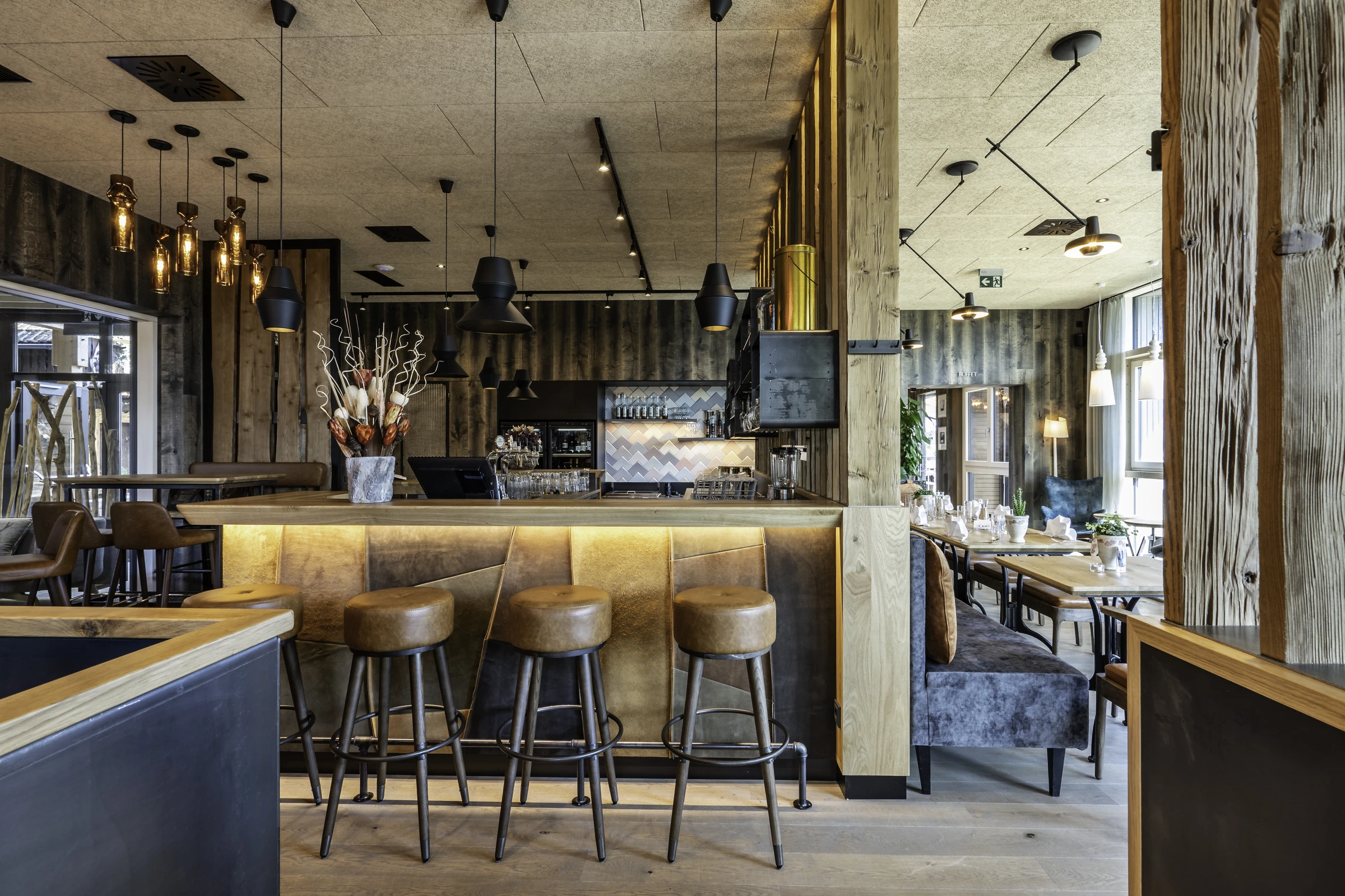 Restaurant und Bar im IKUNA Natternbach mit hinterbeleuchtetem Holztresen, cognacfarbenen Lederbarhockern, schwarzen Industriependelleuchten, Altholzverkleidung und dem Restaurantbereich mit Sitzbänken im Hintergrund.