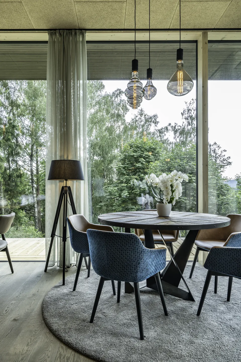 Restaurantbereich im IKUNA Natternbach mit rundem Holztisch, blauen und cognacfarbenen Sesseln, Dreibein-Stehlampe, dekorativen Glühbirnen-Pendelleuchten und großem Panoramafenster mit Waldblick.