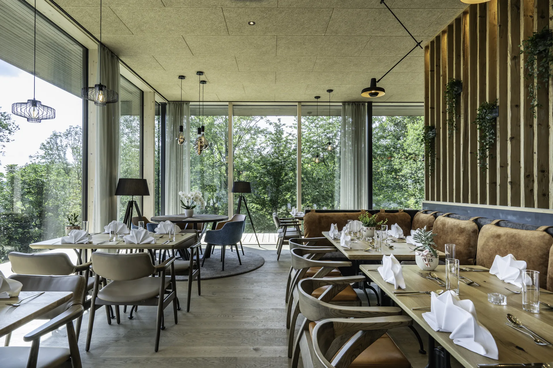 Restaurantbereich im IKUNA Natternbach mit gedeckten Holztischen, verschiedenen Stühlen, cognacfarbener Lederbank, Panoramafenstern mit Waldblick, Stehlampen und einer begrünten Holzlatten-Wand.