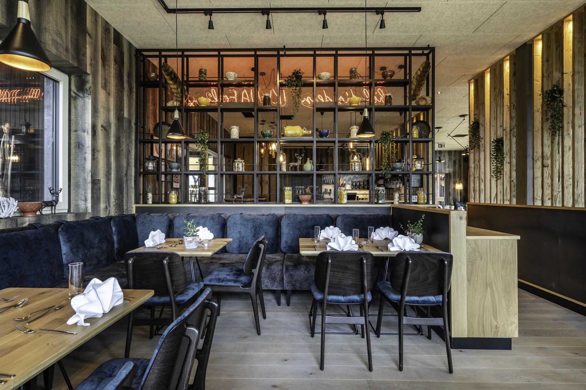 Restaurantbereich im IKUNA Natternbach mit dunkelblauer Eckbank, gedeckten Holztischen, schwarzen Stühlen, einem großen Metallregalraumteiler mit Dekorationen und Neonschrift sowie Altholzverkleidung an den Wänden.