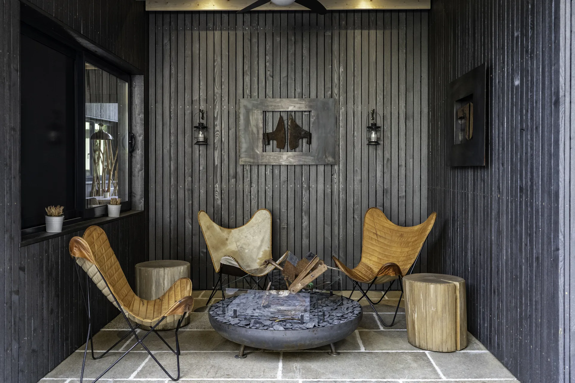 Überdachte Außenlounge im IKUNA Natternbach mit drei Butterfly-Sesseln in Cognac-Leder, einer Feuerschale auf Natursteinboden, Baumstumpf-Beistelltischen und geschwärzter Holzlamellenverkleidung mit Metallkunstwerk an der Wand.