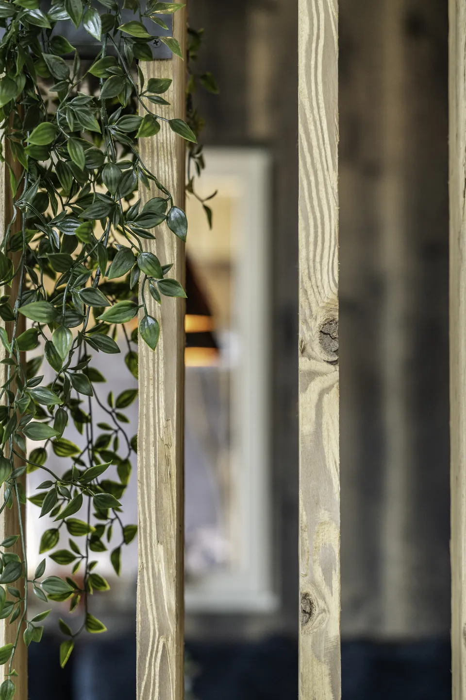 Detailaufnahme im Restaurant des IKUNA Natternbach mit einem Holzlamellen-Raumteiler aus hellem Fichtenholz und einer herabhängenden grünen Rankpflanze, im Hintergrund unscharf der Restaurantbereich.