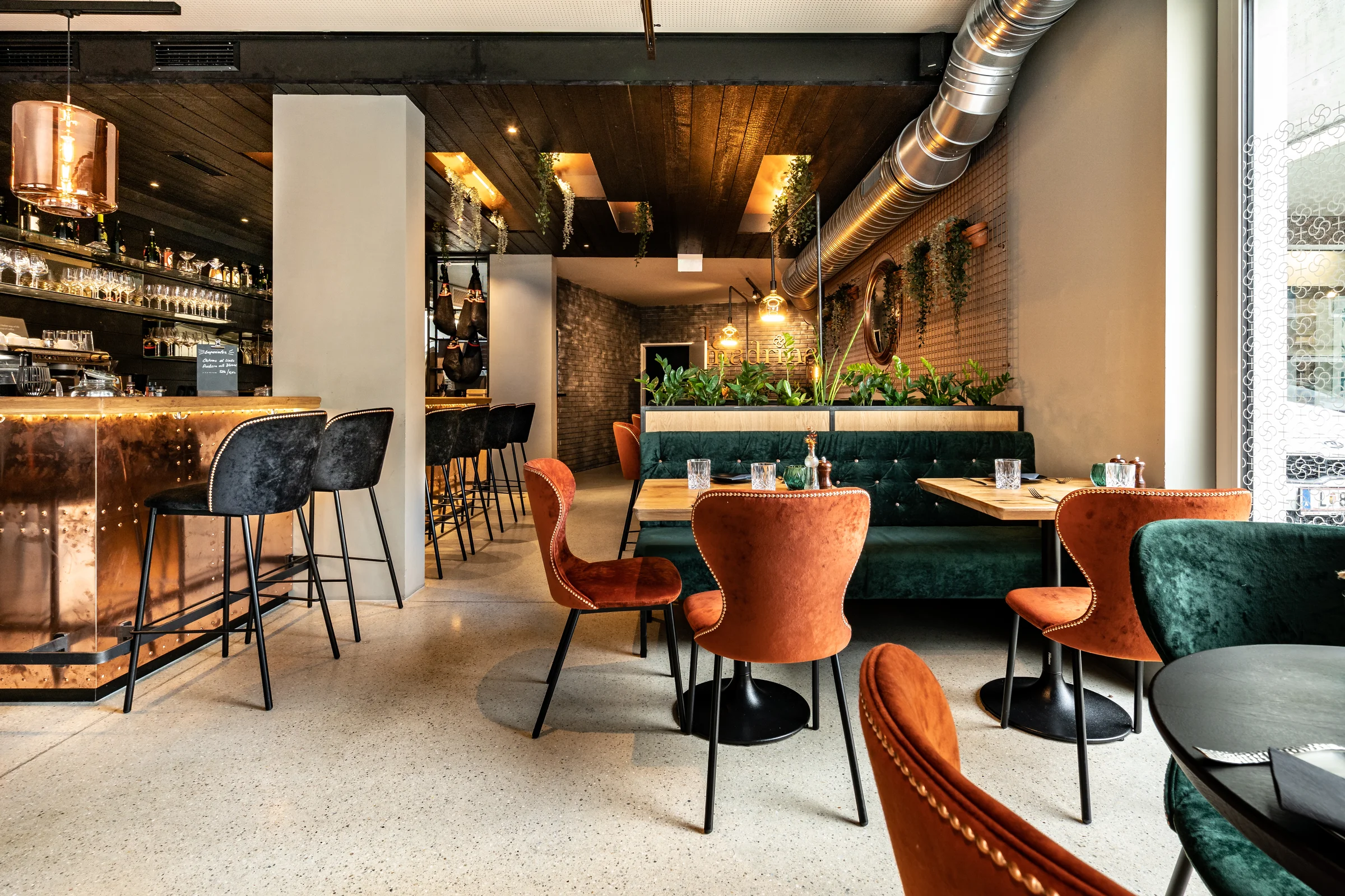 Restaurantbereich La Madrina Linz mit kupferfarbenem Bartresen, schwarzen Barhockern, terrakottafarbenen und dunkelgrünen Samtsesseln mit Nieten, grüner Chesterfield-Sitzbank, Industriedesign-Decke mit Kupferpendelleuchten und begrünter Backsteinwand.