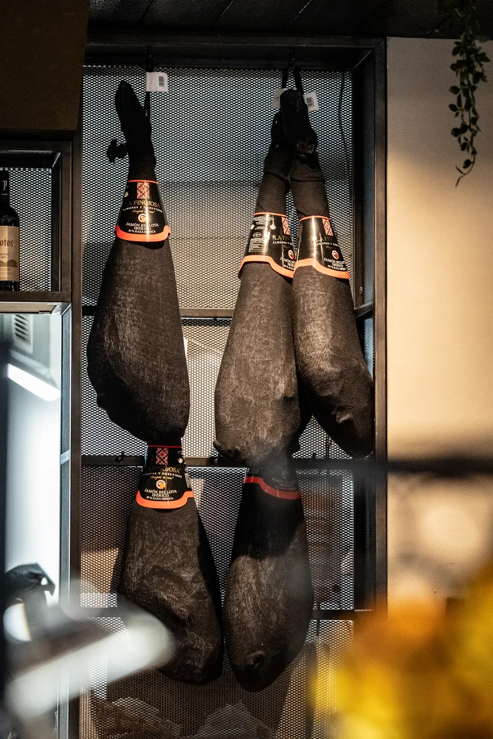 Detailaufnahme im Restaurant La Madrina Linz mit vier hängenden Jamón Ibérico Schinken in schwarzen Schutzhüllen in einem Metallgitter-Regal.