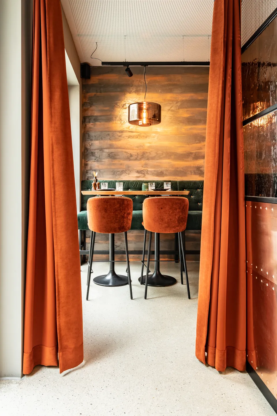 Eingangsbereich im Restaurant La Madrina Linz mit terrakottafarbenen Samtvorhängen, zwei terrakottafarbenen Barhockern, dunkelgrüner Chesterfield-Bank, Kupferpendelleuchte und dekorativer Holzmaserungswand.