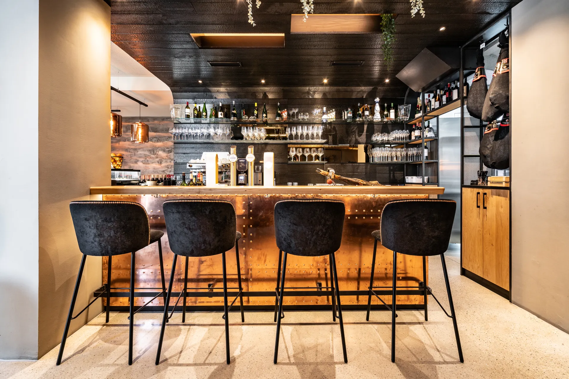 Bar im Restaurant La Madrina Linz mit kupferfarbenem Nietentresen, vier schwarzen Samtbarhockern, hinterbeleuchtetem Weinregal, Kupferpendelleuchten und einem hängenden Jamón Ibérico rechts im Bild.