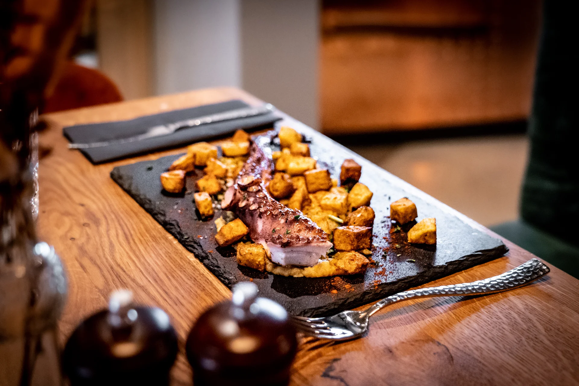 Gegrillter Oktopus mit Patatas Bravas auf einer Schieferplatte, serviert auf einem Holztisch im Restaurant La Madrina Linz, im Vordergrund elegantes Besteck und Pfefferstreuer.