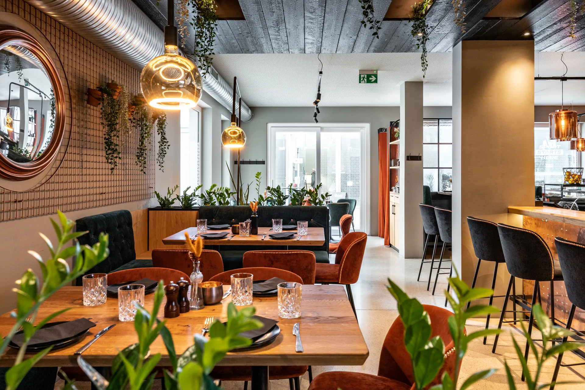 Gastraum im Restaurant La Madrina Linz mit gedeckten Holztischen, dunkelgrüner Chesterfield-Sitzbank, terrakottafarbenen Samtsesseln, goldenen Kugelpendelleuchten, hängenden Rankpflanzen und einem kupferfarbenen Rundspiegel.