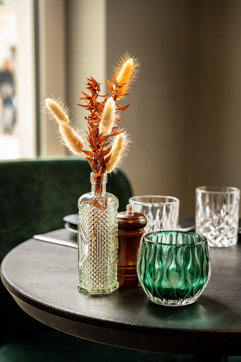Tischdetail im Restaurant La Madrina Linz mit einer Glasvase mit Trockenblumen und Pampasgras, einem Holzpfefferstreuer, grünem Teelichtglas und Kristallgläsern auf einem dunklen Rundtisch vor grüner Samtbank.