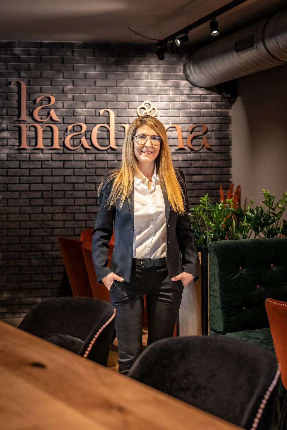 Inhaberin des Restaurants La Madrina Linz lächelnd vor der dunklen Backsteinwand mit kupferfarbenem La-Madrina-Schriftzug, im Hintergrund grüne Samtsessel und Zimmerpflanzen.
