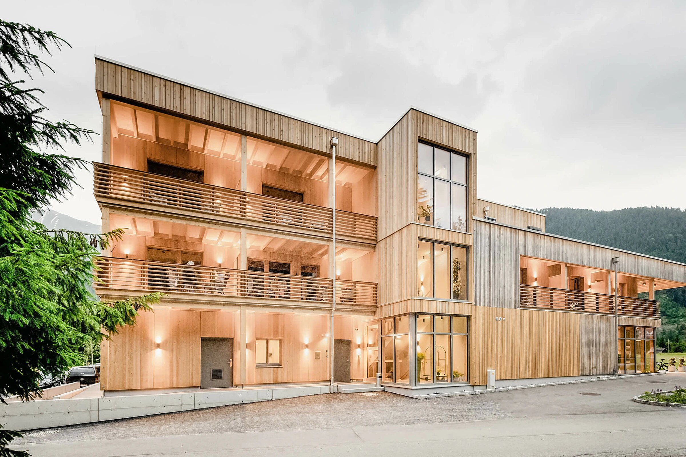 Außenfassade des LechZeit in Elmen mit naturbelassener Holzverkleidung, warm beleuchteten Balkonen mit Holzgeländern und Bergpanorama im Hintergrund.