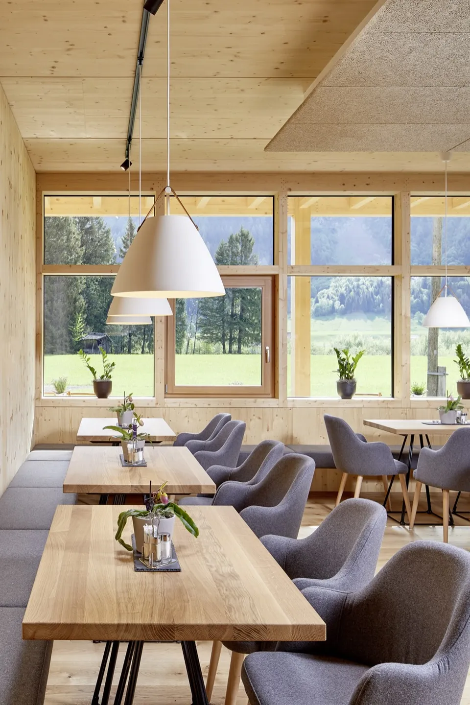 Frühstücksraum des LechZeit in Elmen mit heller Holzvertäfelung, grauen Polstersesseln, Naturholztischen, weißen Pendelleuchten und Panoramafenstern mit Blick auf die grüne Bergwiese.