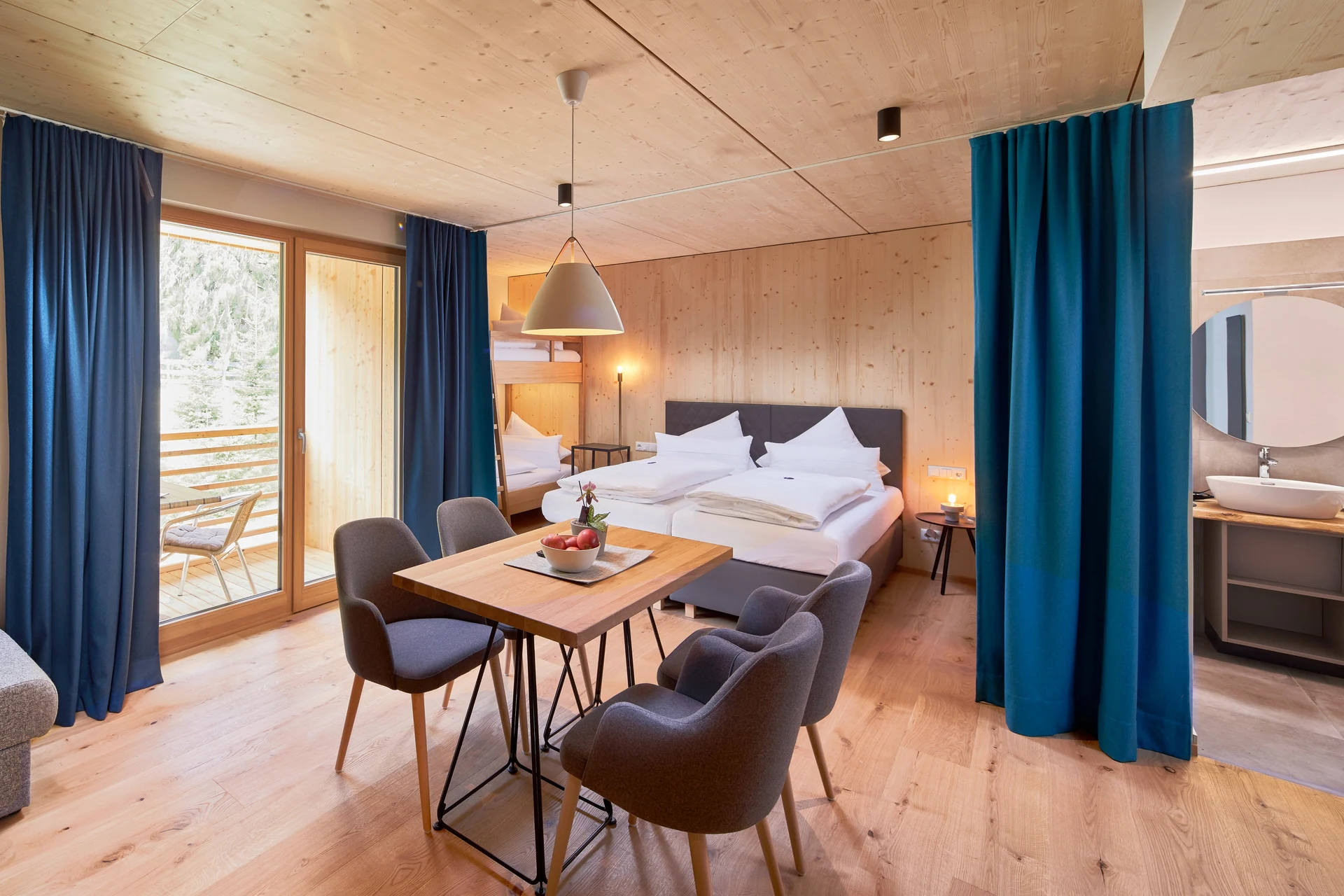 Suite des LechZeit in Elmen mit Wohnbereich, Esstisch mit grauen Sesseln, Doppelbett, blauen Vorhängen, Balkon mit Waldblick, Holzdecke und offenem Badbereich mit Spiegel.