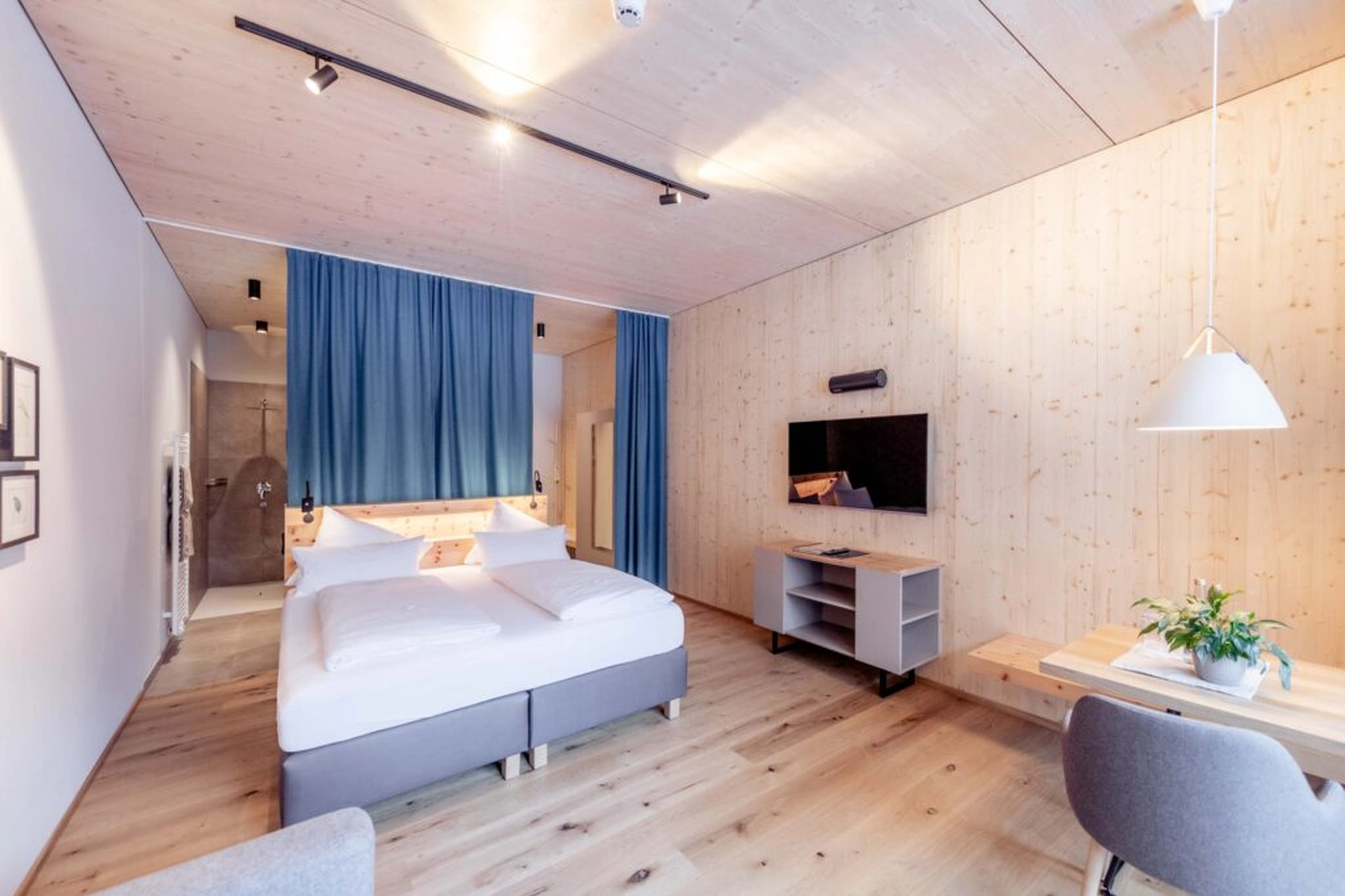 Hotelzimmer des LechZeit in Elmen mit Doppelbett, heller Holzvertäfelung, blauem Vorhang als Raumtrenner zum offenen Badbereich, Fernseher, Sideboard und Schreibtisch mit Pendelleuchte.