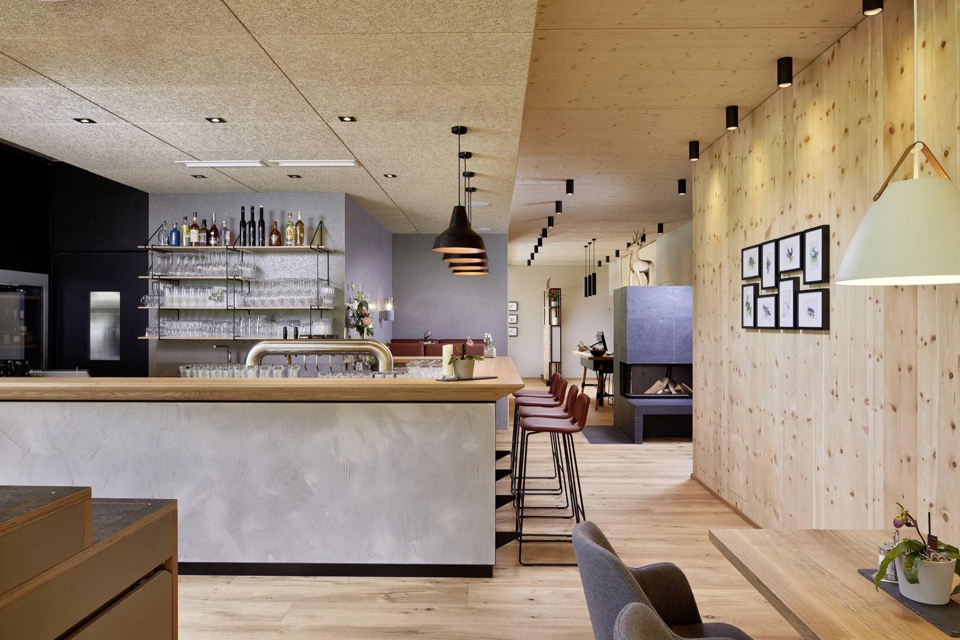 Bar des LechZeit in Elmen mit hellem Holz-Beton-Tresen, roten Barhockern, goldenem Glasregal, schwarzen Pendelleuchten, modernem Kamin, Hirschskulptur und naturbelassener Holzwand mit Tierbild-Galerie.